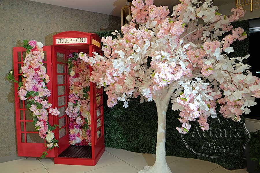 Фотозона "Телефонная будка" с цветами и бело-розовым деревом на фоне стены из самшита
